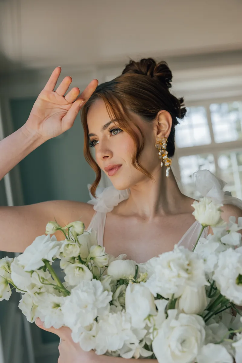 Mariée au chignon, portant des boucles florales dorées et tenant un bouquet blanc